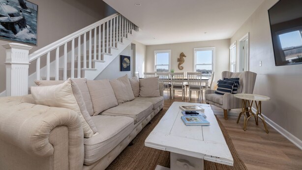 A spacious living room with a beige sectional sofa, a white coffee table, and a dining area with white chairs. A staircase with white railings leads to an upper level. - Amazing 10 Bedroom Compound on the Water with 6 Bedroom Home & 4 Studio Cottages (Stonington)