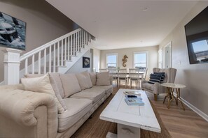 A spacious living room with a beige sectional sofa, a white coffee table, and a dining area with white chairs. A staircase with white railings leads to an upper level.