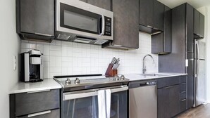 A modern kitchen with dark cabinetry, a built-in microwave, a coffee machine, a sink, and a dishwasher. - Landing | Beautiful Studio, Courtyard, Yoga Studio (Durham)