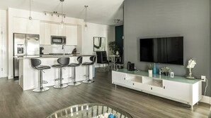 A modern kitchen with a central island, bar stools, a large flat-screen TV, and a minimalist design. - Landing | Modern 2BD, Gym, Pool (Tampa)