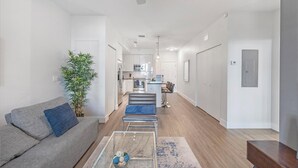 A modern living room with a grey sofa, a coffee table, and a plant. A kitchen with white cabinets and stainless steel appliances is visible in the background. - Landing | Amazing 1BD, Clubhouse, Pool (Tampa)