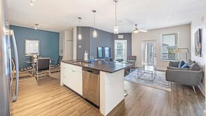 A modern kitchen with a central island, a dining area with a glass table and chairs, a gray sofa, and a large window with blinds. - Landing | Modern 2BD, Clubhouse, Gym (Tampa)
