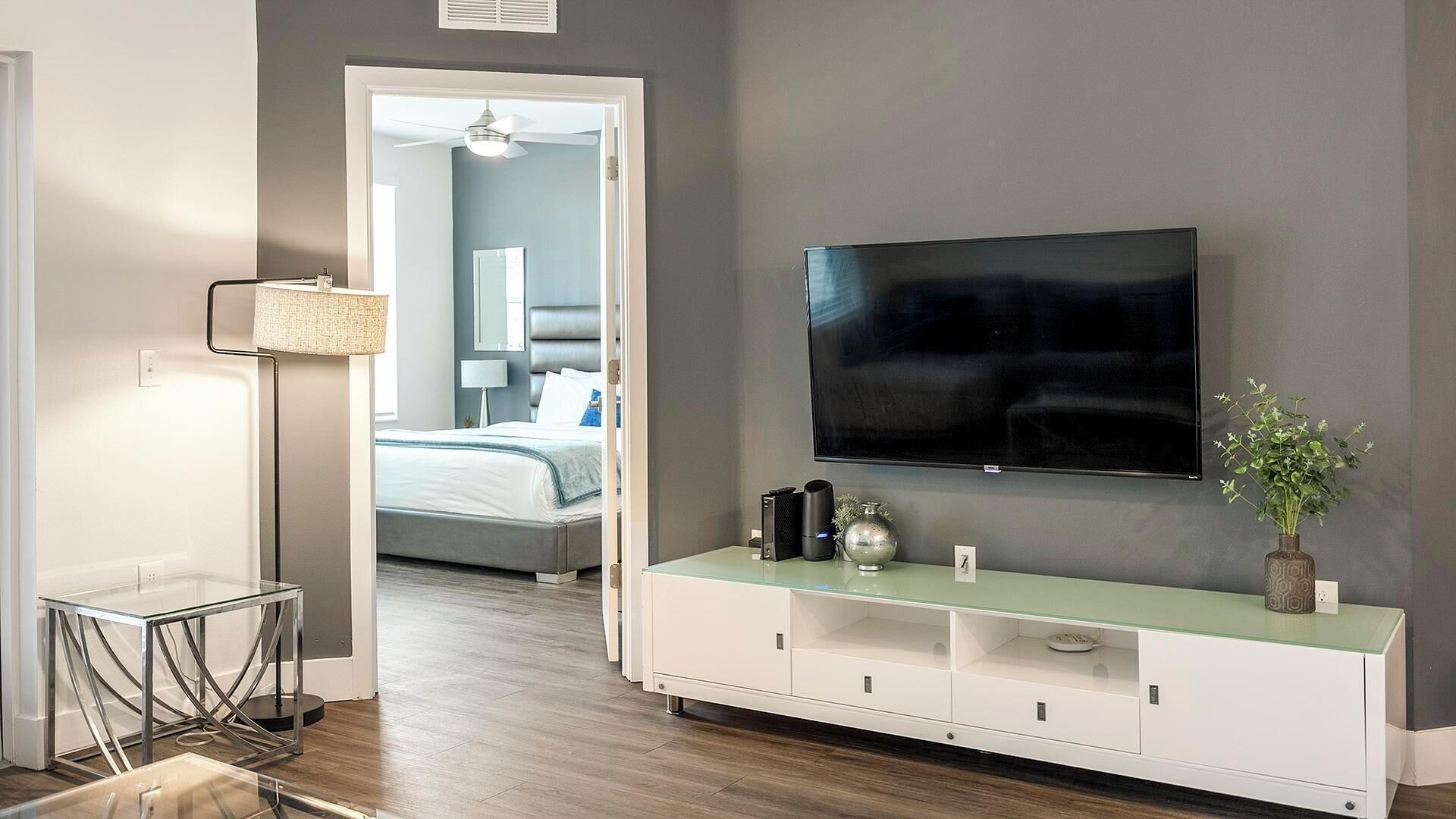 Una habitación de hotel moderna con un televisor de pantalla plana grande montado en la pared, una mesita de centro con cubierta de vidrio y objetos decorativos, y vista al dormitorio contiguo a través de un umbral abierto.