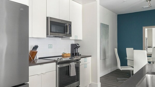 A modern kitchen with white cabinetry, a stainless steel refrigerator, a microwave, and a coffee machine. There is a dining area with a table and chairs.