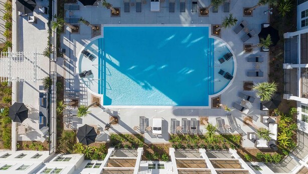 Ein Luftbild eines modernen Hotels mit einem zentral gelegenen Schwimmbecken, umgeben von Liegestühlen und einer überdachten Terrasse.