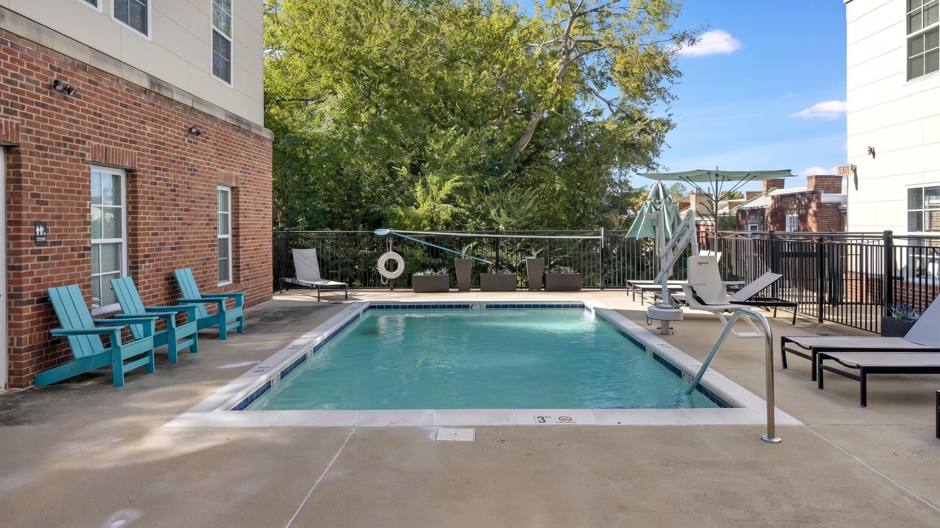 A rectangular pool with a safety ladder, surrounded by a concrete deck. There are lounge chairs and a small table with an umbrella.