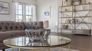 A living room with a glass coffee table, a brown sofa, a bookshelf, and a framed picture on the wall.