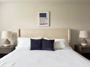 Un lit bien fait, avec des oreillers blancs et bleu foncé, une tête de lit beige et un tableau encadré au-dessus.