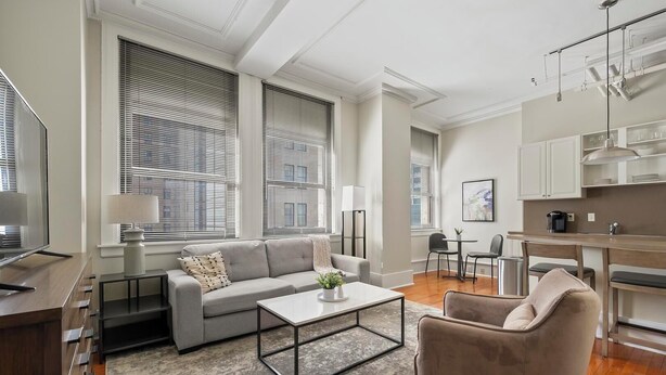Una sala moderna con un sofá gris, una mesa de centro y un sillón beige. Se observa una zona de cocina con una mesa de comedor y sillas, y ventanas amplias con persianas.