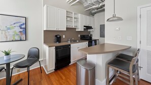 Una cocina moderna con gabinetes blancos, lavavajillas, estufa y fregadero. Hay un comedor con una mesa y sillas, además de un cubo de basura.