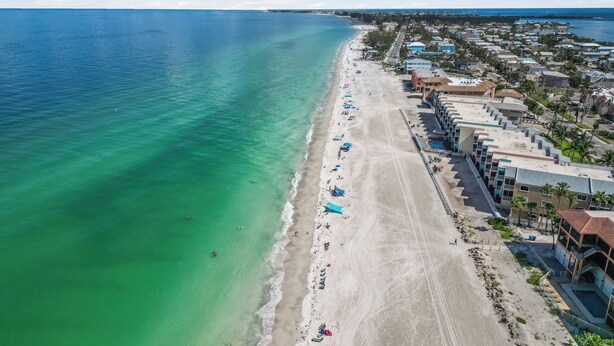 Vue aérienne d’un complexe hôtelier situé en bord de mer, avec une longue plage de sable, des eaux turquoise et limpides, et une rangée d’immeubles à plusieurs étages dotés de balcons.