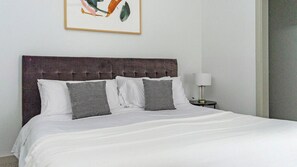 Una cama bien hecha con ropa de cama blanca y gris, un cabecero oscuro y una mesita de noche pequeña con una lámpara.