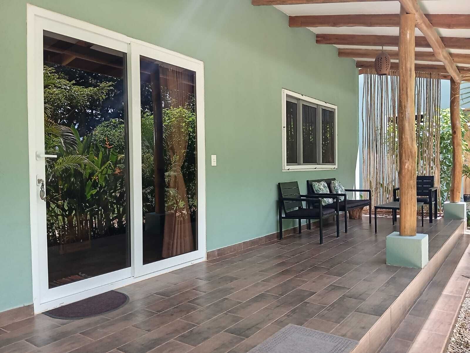 A covered patio area with a sliding glass door leading to a lush garden, a seating area with chairs and a table, and a wooden pergola structure.