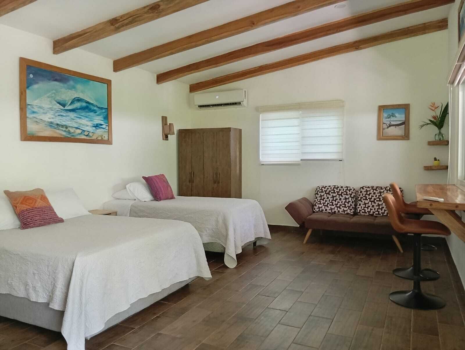 Ett rum med två enkelsängar, en soffa, ett träpanelat tak och ett fönster med persienner.