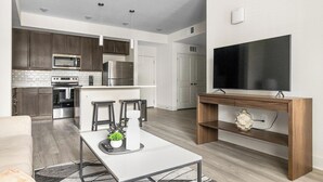 A modern kitchen with stainless steel appliances, a dining area with a white table and bar stools, and a large flat-screen TV mounted on a wooden console.