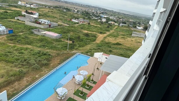 A view of a swimming pool with lounge chairs and umbrellas, surrounded by greenery and buildings in the background.
