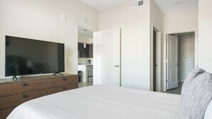 A bedroom with a large bed, a TV on a dresser, and a view of a kitchen and bathroom through an open door.