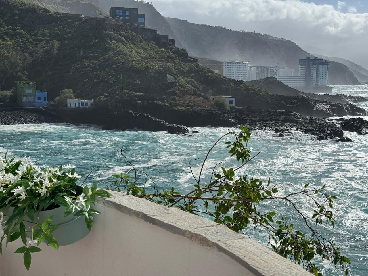 Vista costera con costa rocosa, olas rompiendo contra las rocas y edificios al fondo.