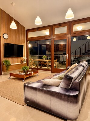 A modern living room with a leather sofa, wooden panel walls, a flat-screen TV, and a dining area with a glass door.