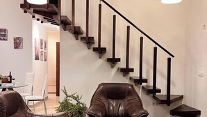 Una sala de estar moderna con un sofá de cuero marrón, un sillón de cuero y una mesa de centro de mármol. Una escalera de madera con barandillas negras conduce a un nivel superior.