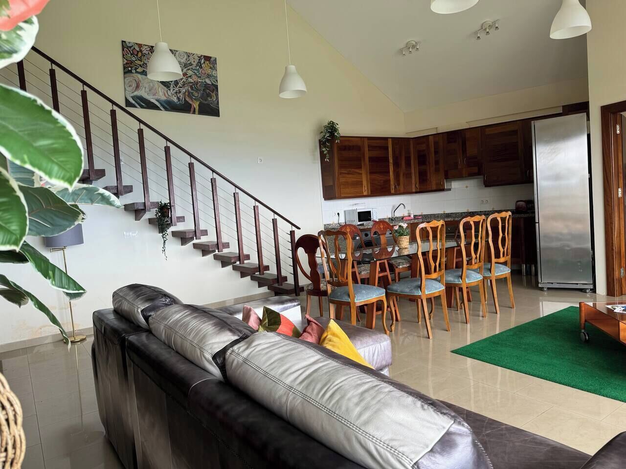 A spacious living area with a leather sofa, dining table with chairs, wooden cabinets, and a staircase.