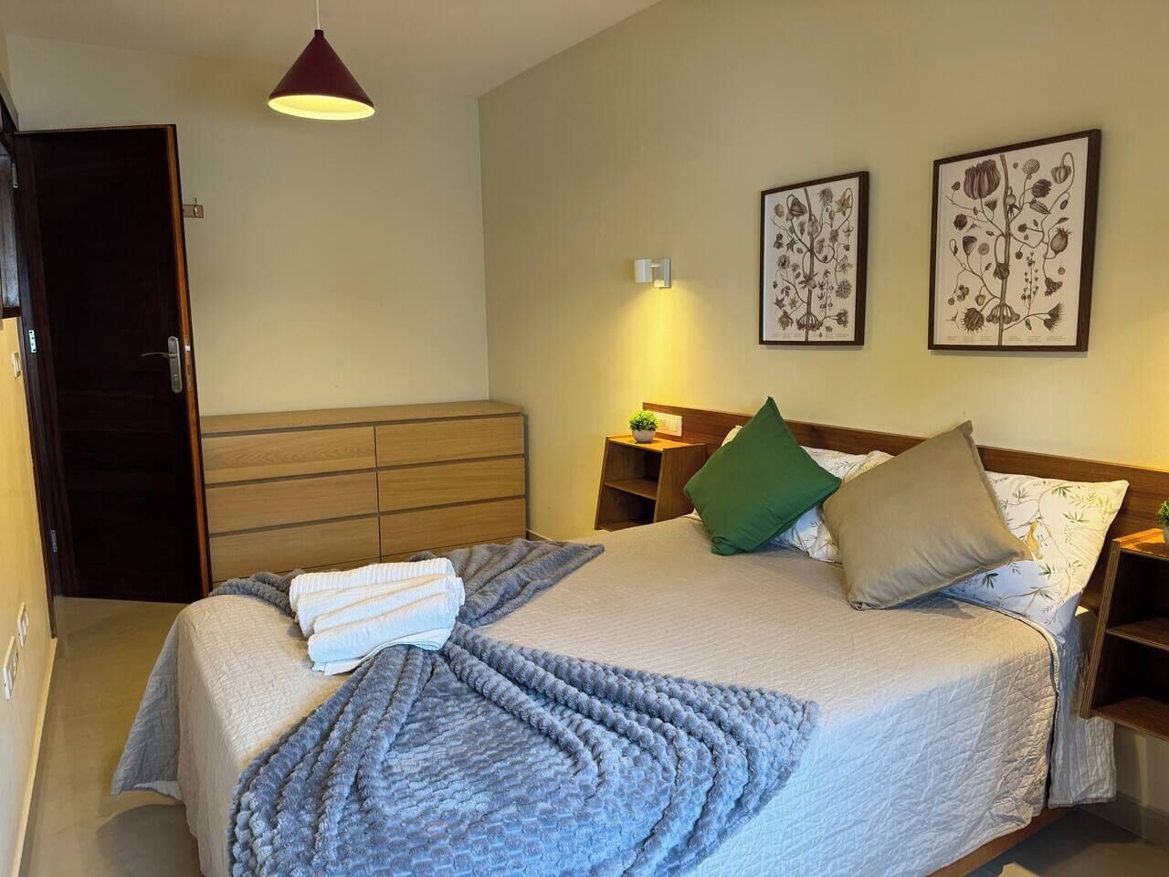 Una cama bien hecha con una manta gris y almohadas, una mesita de noche con una lámpara, y dos cuadros con motivos florales en la pared.