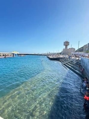A clear waterfront with a pier extending into the water, a building with a tower, and people swimming.
