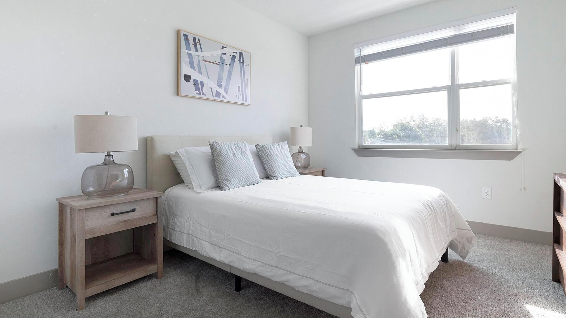 A bedroom with a bed, two nightstands, a window, and a wall-mounted abstract painting.