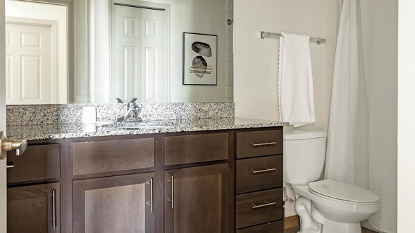 Un baño con armarios de madera oscura, encimera de granito, inodoro y cortina de ducha blancos.
