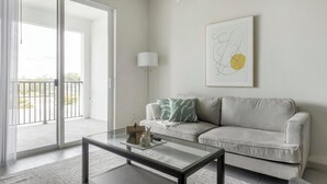 Una sala de estar moderna con una mesa de centro de cristal, un sofá gris con cojines estampados, una lámpara de pie y una obra de arte abstracto en la pared.