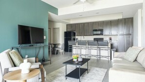 Una sala moderna con televisor, sofá y mesa de centro. Se ve una cocina con armarios oscuros y microondas al fondo.