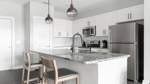 A modern kitchen with white cabinets, a stainless steel refrigerator, a microwave, and a granite countertop. There are two wooden chairs and a pendant light above the island.