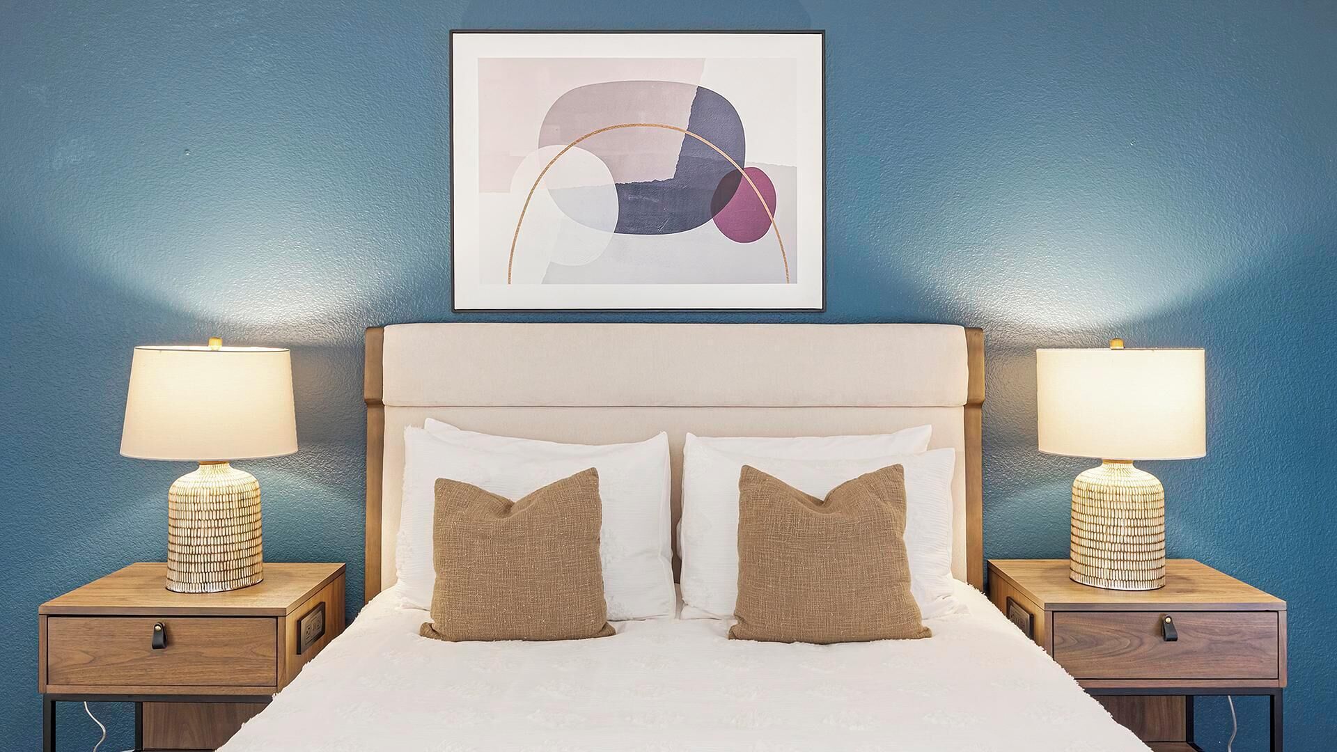 Ein Schlafzimmer mit einem Bett, zwei Nachttischen, zwei Lampen und einem gerahmten abstrakten Kunstwerk an der Wand.