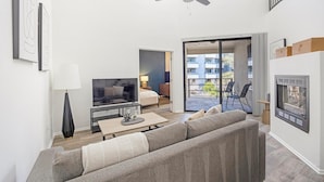 A modern living room with a sofa, TV, and a dining area. There is a balcony with a view of buildings and a door leading outside.