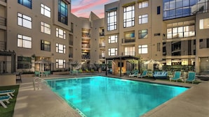 A modern hotel with a central swimming pool, outdoor seating, and a multi-story building with balconies.