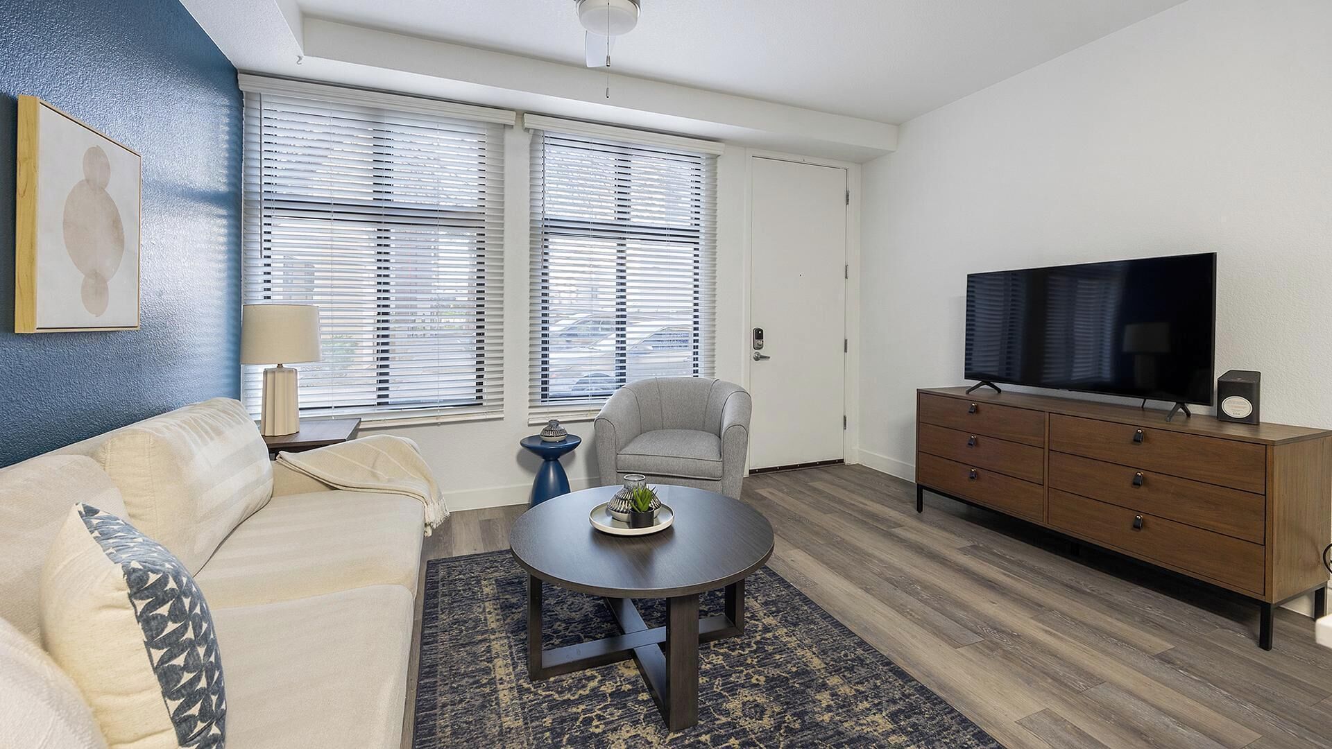 A living room with a sofa, a round coffee table, a grey armchair, a TV on a wooden cabinet, and a window with blinds.