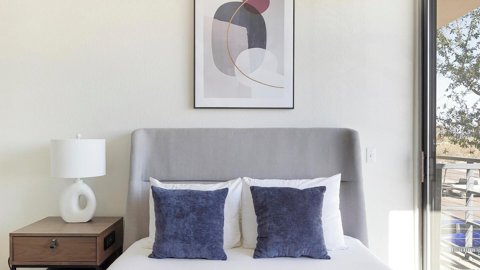 Een hotelkamer met een bed, nachtkastje, lamp en een lijst met abstracte kunst aan de muur.