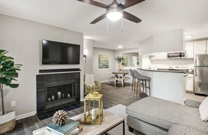 A modern living room with a fireplace, a dining area, and a kitchen with white cabinets and stainless steel appliances.