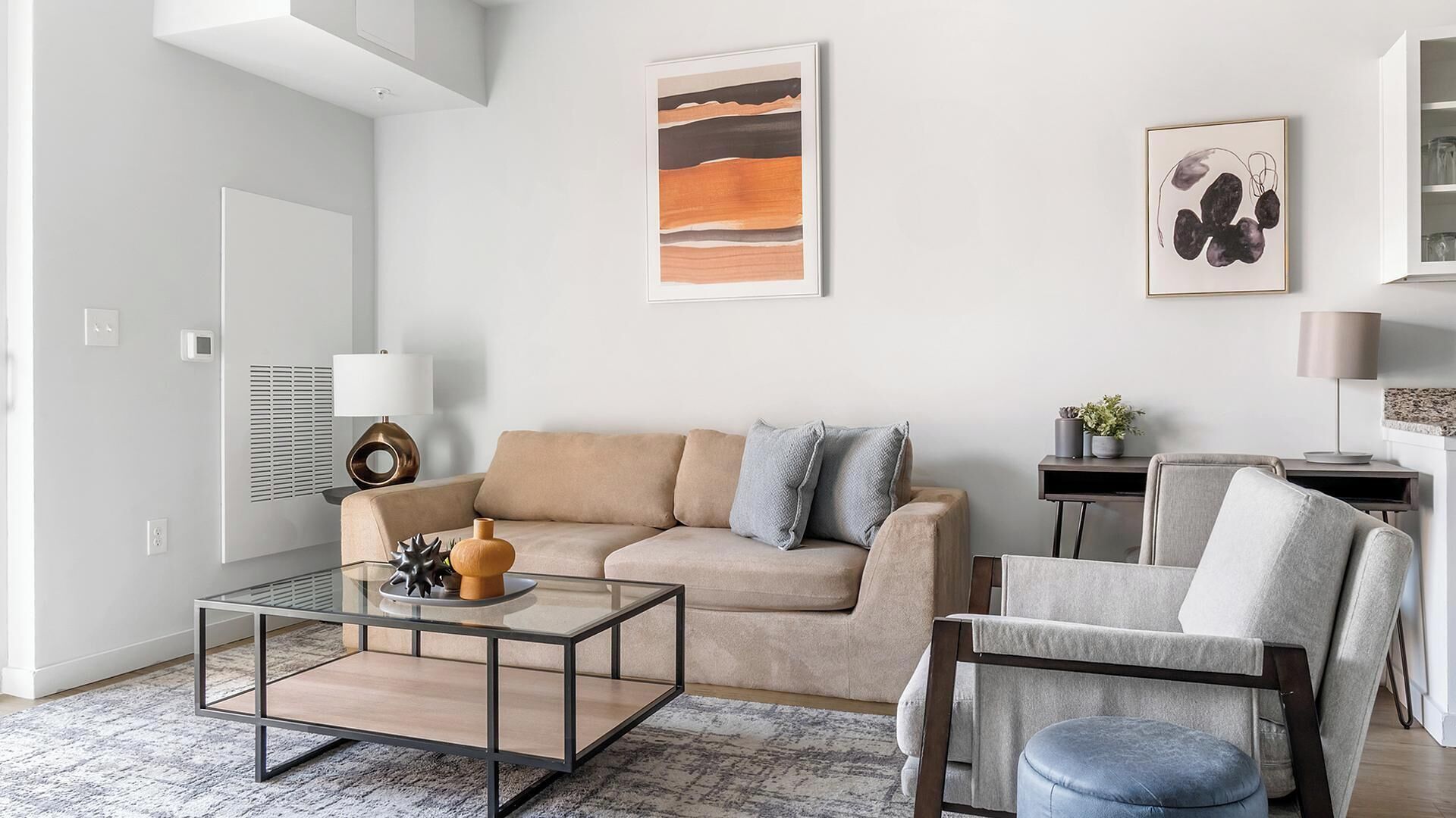 Un salon moderne avec un canapé beige, une table basse en verre et un fauteuil gris. Un tableau est accroché au mur et une lampe se trouve sur une table d’appoint.