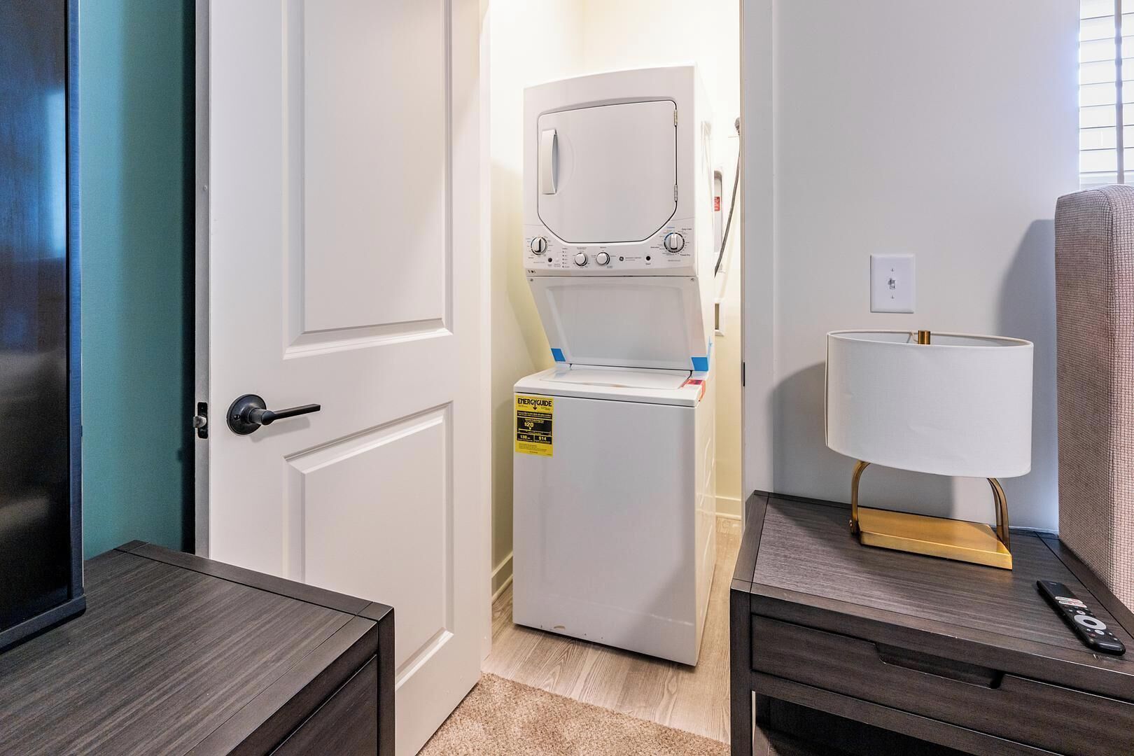 Une chambre d’hôtel avec une porte blanche, un lave-linge, une lampe et une petite table avec une télécommande.
