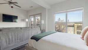 A bedroom with a large bed, a ceiling fan, a TV mounted on the wall, and a window with a city view.
