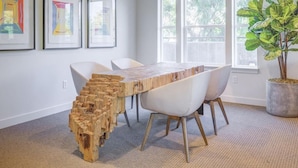 A modern dining area with a unique wooden bench, white chairs, and a large window with a view of trees.