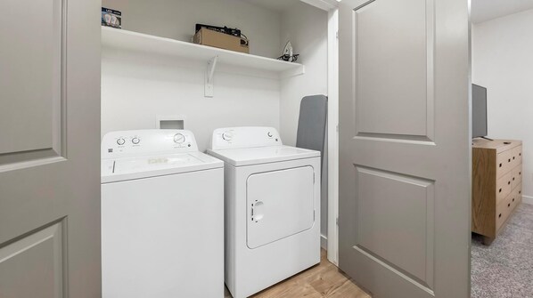 A laundry area with two white washing machines and a dryer, a wooden cabinet, and a shelf with items.