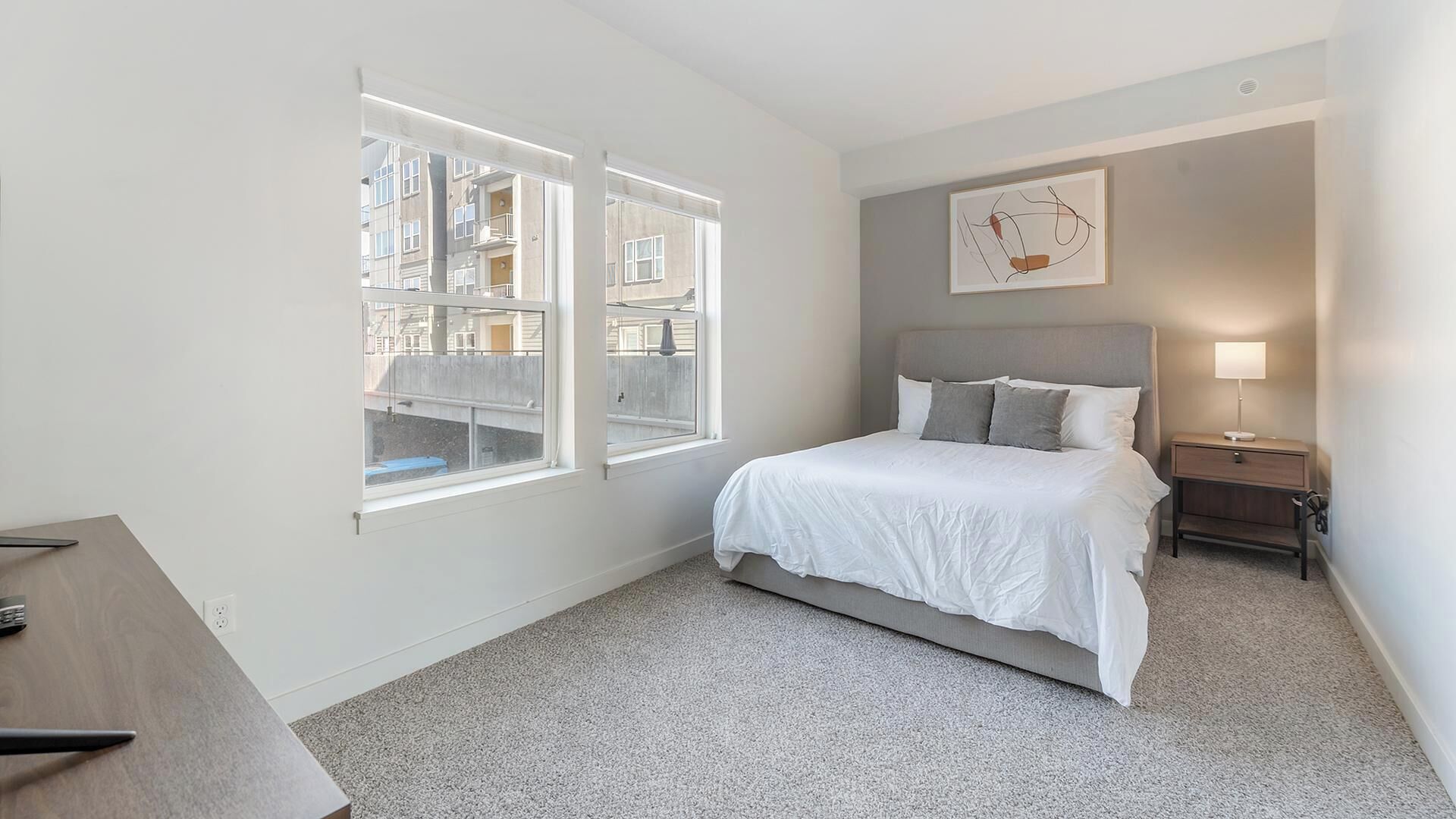 Un dormitorio con cama, mesita de noche, lámpara y ventana con vistas a edificios.