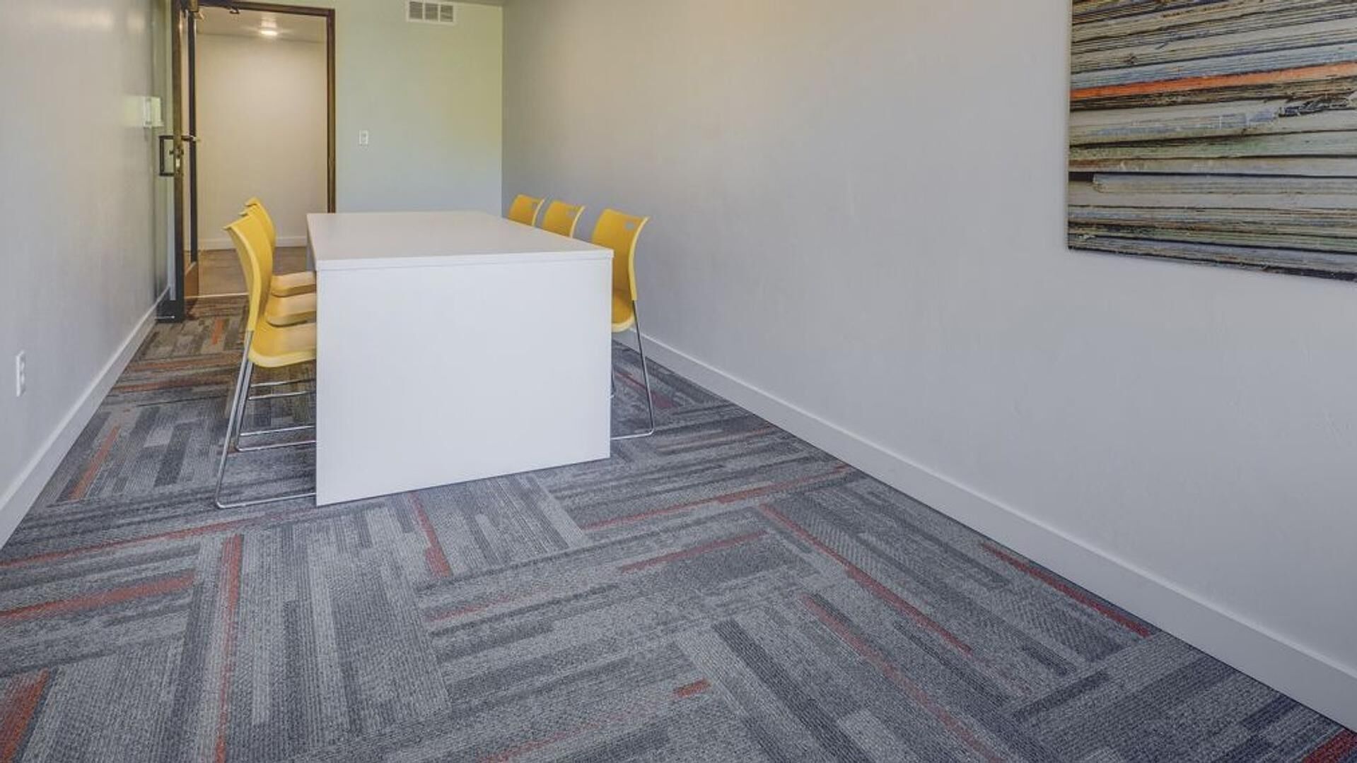 A conference room with a white table and yellow chairs, carpeted floor, and a plain wall with a textured artwork.