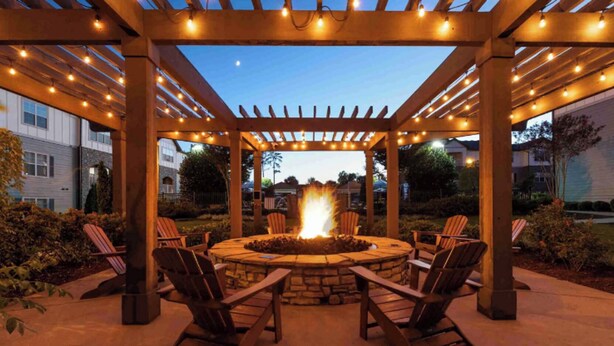 Un espacio al aire libre con asientos circulares, una chimenea rodeada de sillas de madera y una pérgola con luces colgantes.