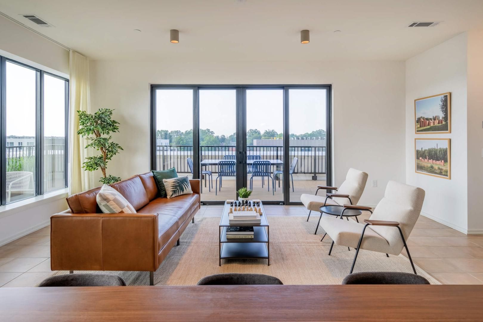 Un salon moderne avec un canapé en cuir, une table basse sur laquelle sont posés des livres et une vue sur un balcon aménagé avec du mobilier d’extérieur.