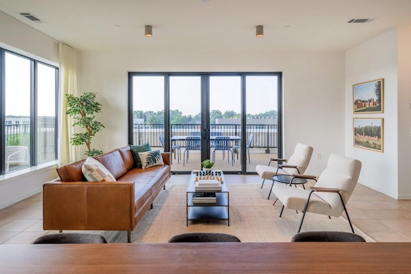 A modern living room with a leather sofa, a coffee table with books, and a view of a balcony with outdoor furniture.