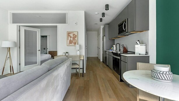 A modern hotel room with a bed, bedside table, lamp, and a view of the kitchen.