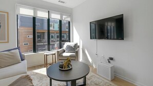A modern living room with a large window, a flat-screen TV mounted on the wall, a round coffee table, and a comfortable seating area with a sofa and armchair.